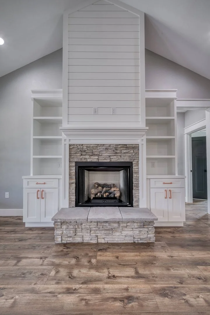 Interior view of a living room fireplace with a stone facade, flanked by white built-in shelves and cabinets, with a wood floor and a white wall and ceiling.