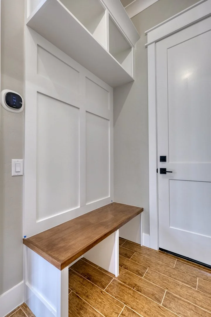 Small entry nook with white paneled walls, a wooden bench, white shelving above, and a white door with black hardware.
