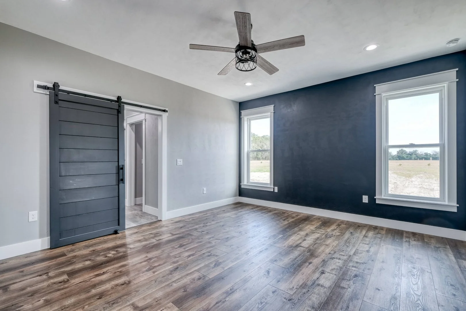 Empty room with two large windows, blue accent wall, wood flooring, ceiling fan, and a sliding barn door leading to an adjacent room.