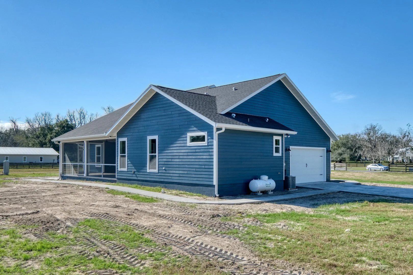 A blue house with a gray roof, a white garage door, small windows, and a screened-in porch, situated on a grassy and partly dirt yard under a clear blue sky.