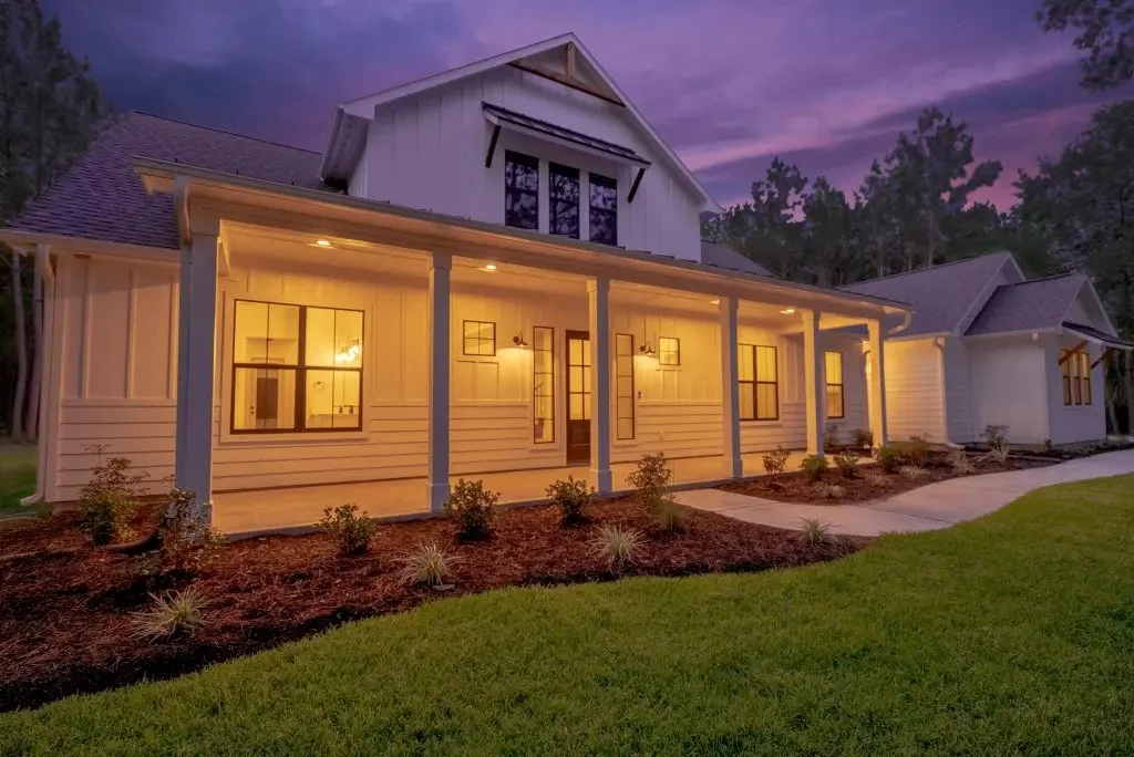 Exterior view of a two-story house at dusk with lit windows, a porch with pillars, and a landscaped yard with small bushes and grass.