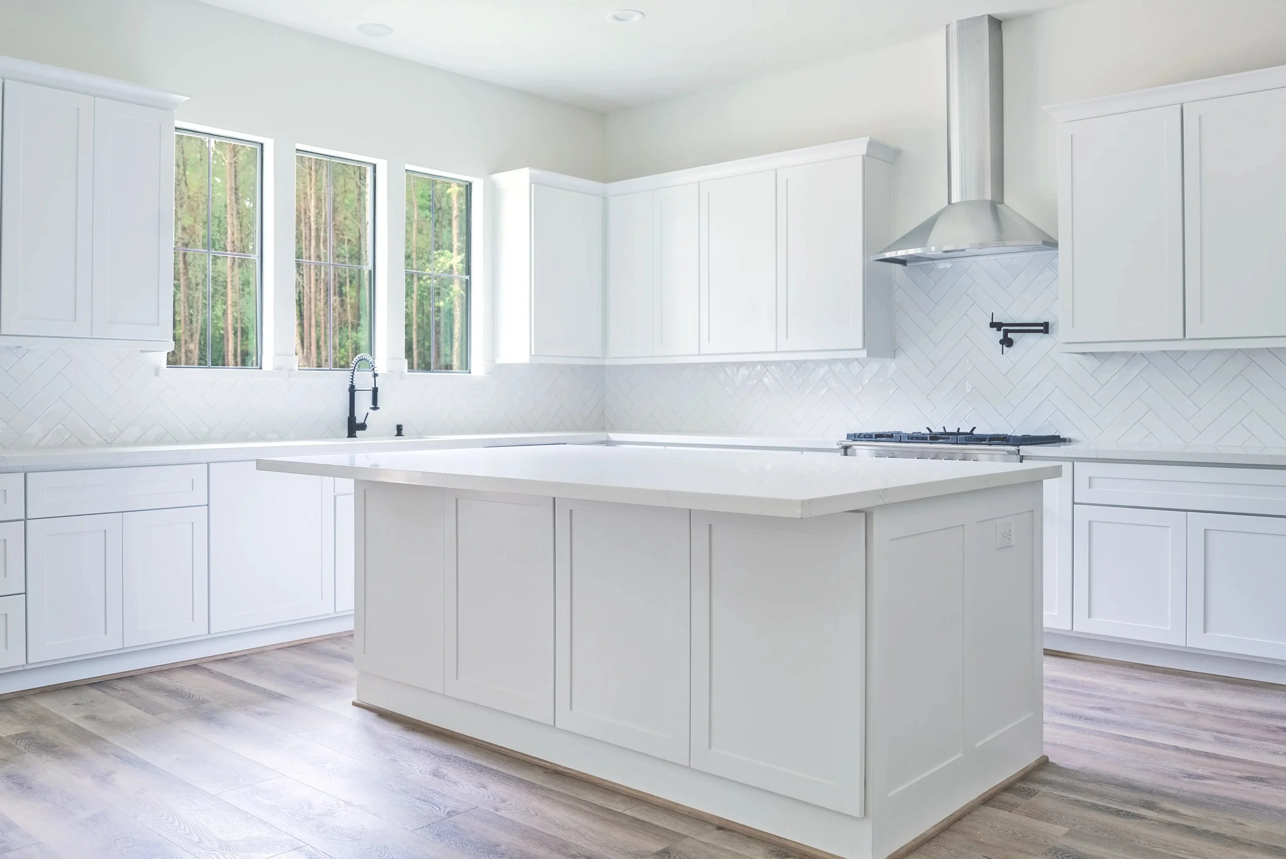 Bright, modern kitchen with white cabinets, a white island, a herringbone patterned white backsplash, a stainless steel range hood, black faucet, and large windows overlooking trees.