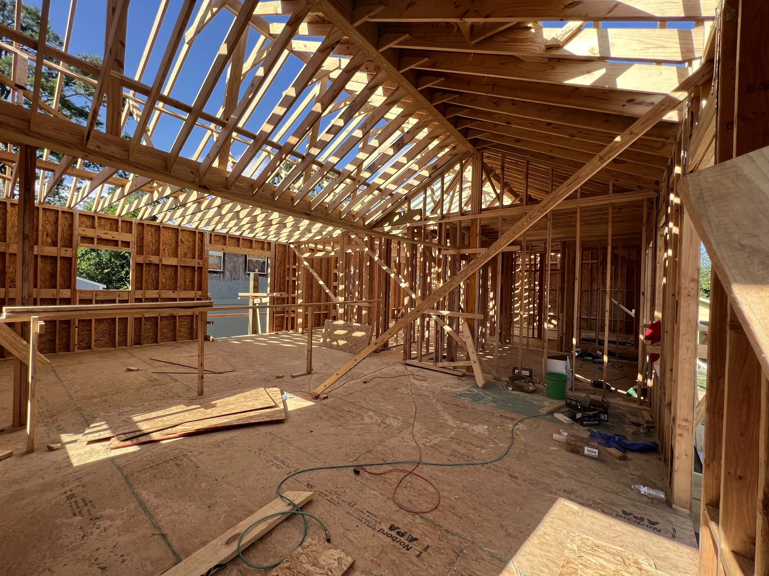 Interior view of a house under construction with exposed wooden framing, roof trusses, and unfinished walls and floor.
