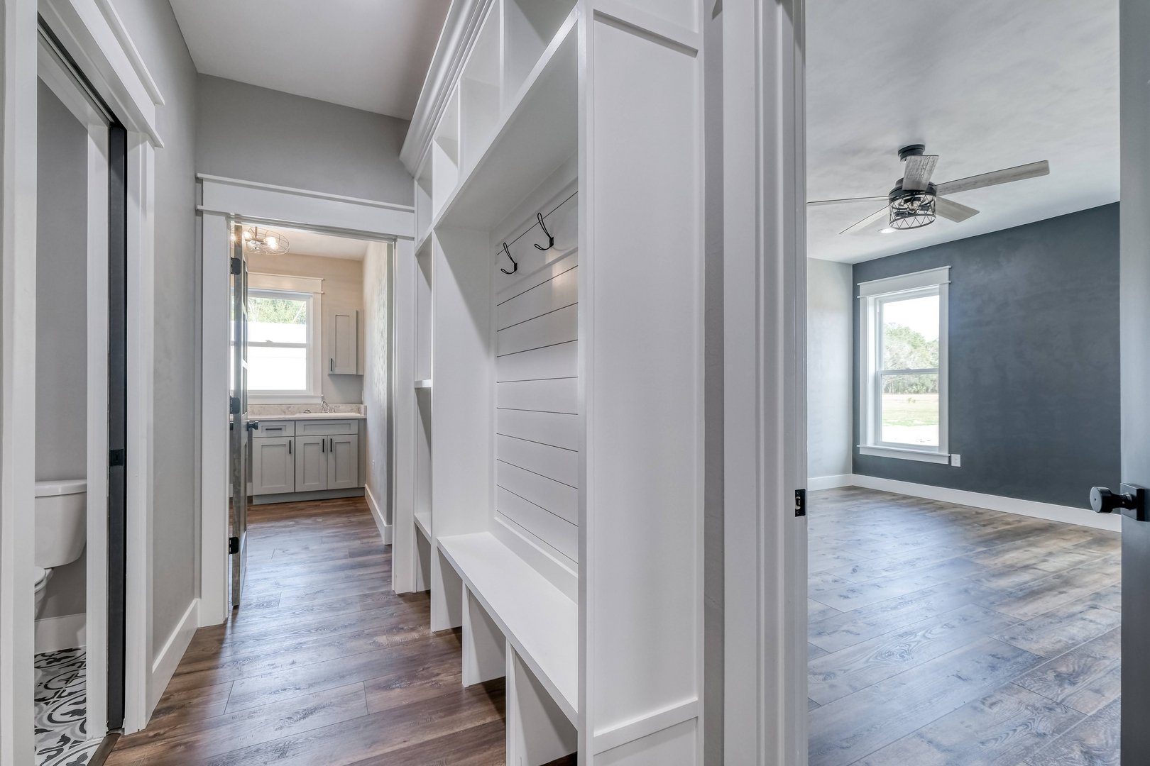 Interior view of a modern house with a laundry area on the left and a bedroom on the right, featuring hardwood floors, white and gray walls, and large windows.