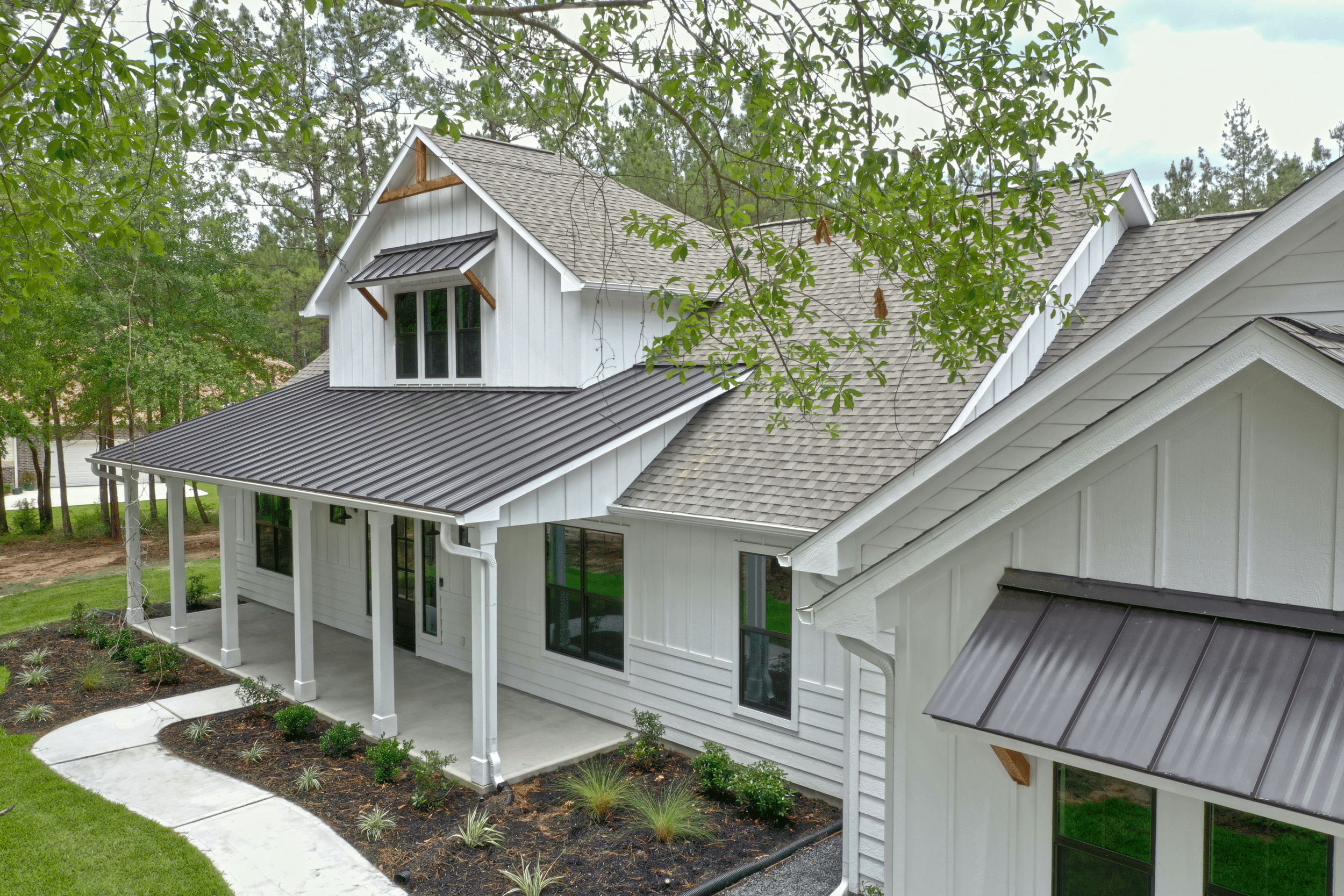 White two-story house with gray roof, front porch, and landscaped yard with small bushes and a curved concrete walkway, surrounded by trees.