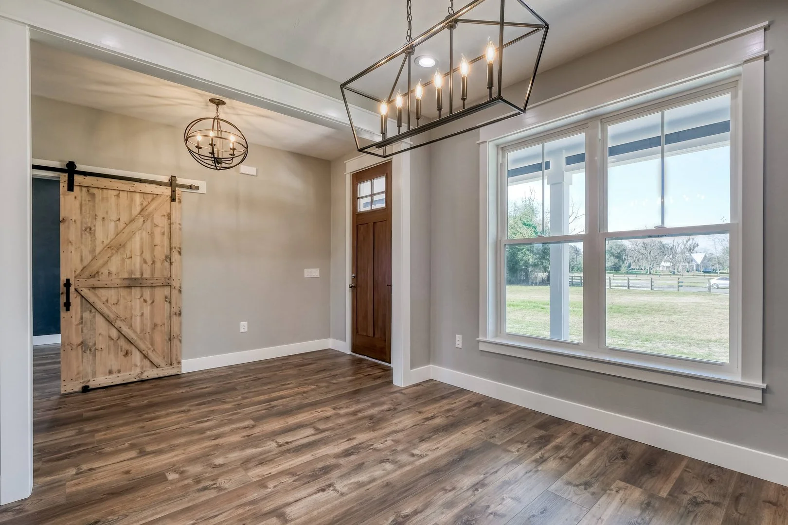 Empty room with large window, wooden door, wood floor, two modern light fixtures, and sliding barn door.