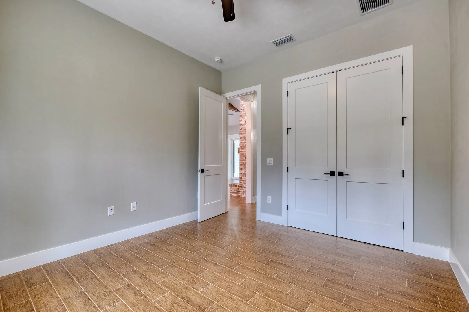 Empty room with light gray walls, wooden flooring, an open door leading to another room with brick wall, and a large white double closet with black handles.