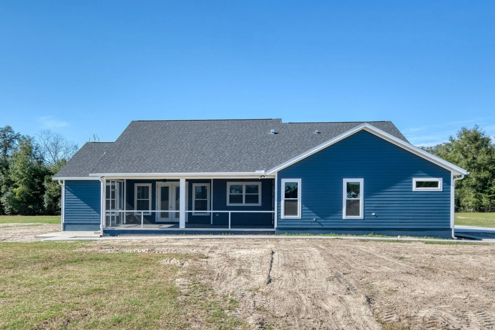 New blue house with a large front porch, sliding glass doors, multiple windows, and a gray roof, set on a grassy plot with a dirt pathway in front and trees in the background under a clear blue sky.