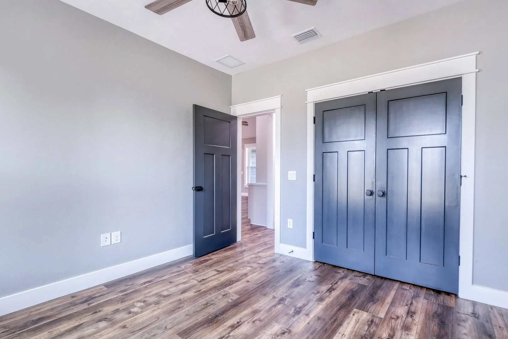 Empty room with hardwood floors, gray doors, and white trim, with a ceiling fan and vents.