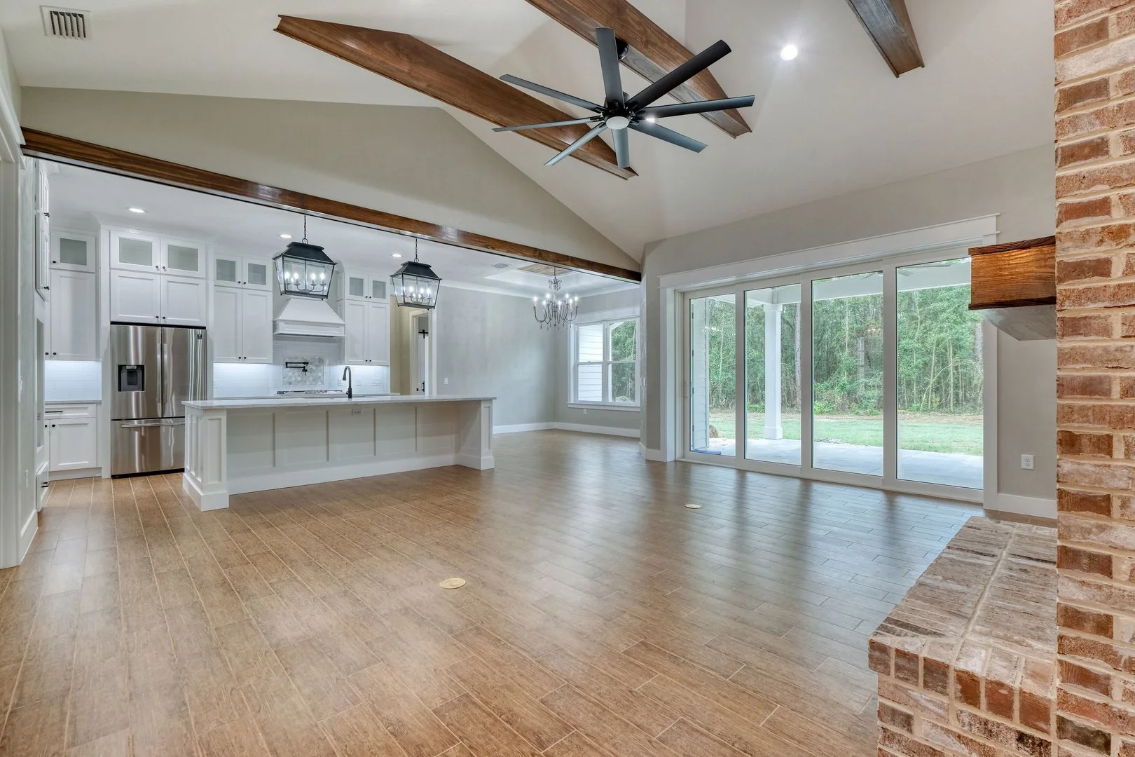 Open concept living area with kitchen, large sliding glass doors, wood flooring, brick fireplace hearth, exposed wooden beams, and ceiling fan.