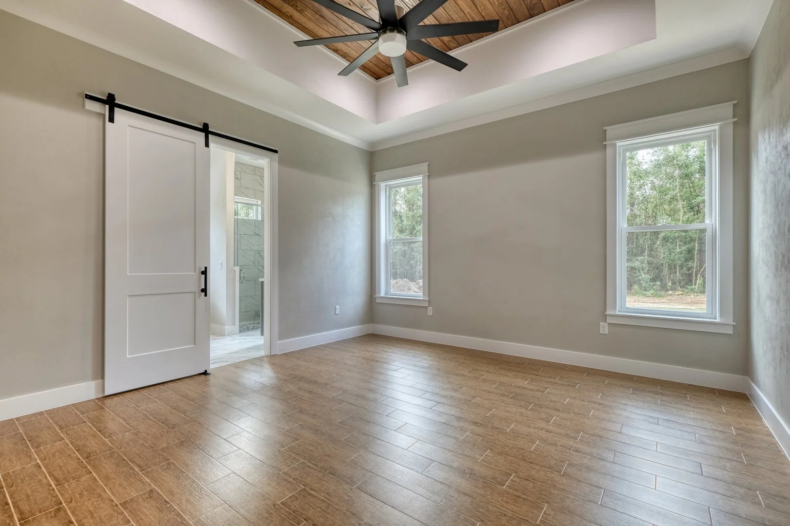 Empty bedroom with wood flooring, two windows, a ceiling fan, and a sliding barn door leading to a bathroom.