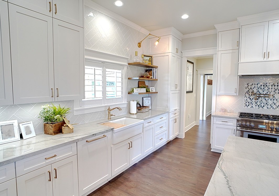 Modern white kitchen with wood flooring, white cabinets, a window with shutters, and open shelves with decor and books. Contains a farmhouse sink, a stovetop, and a marble countertop.