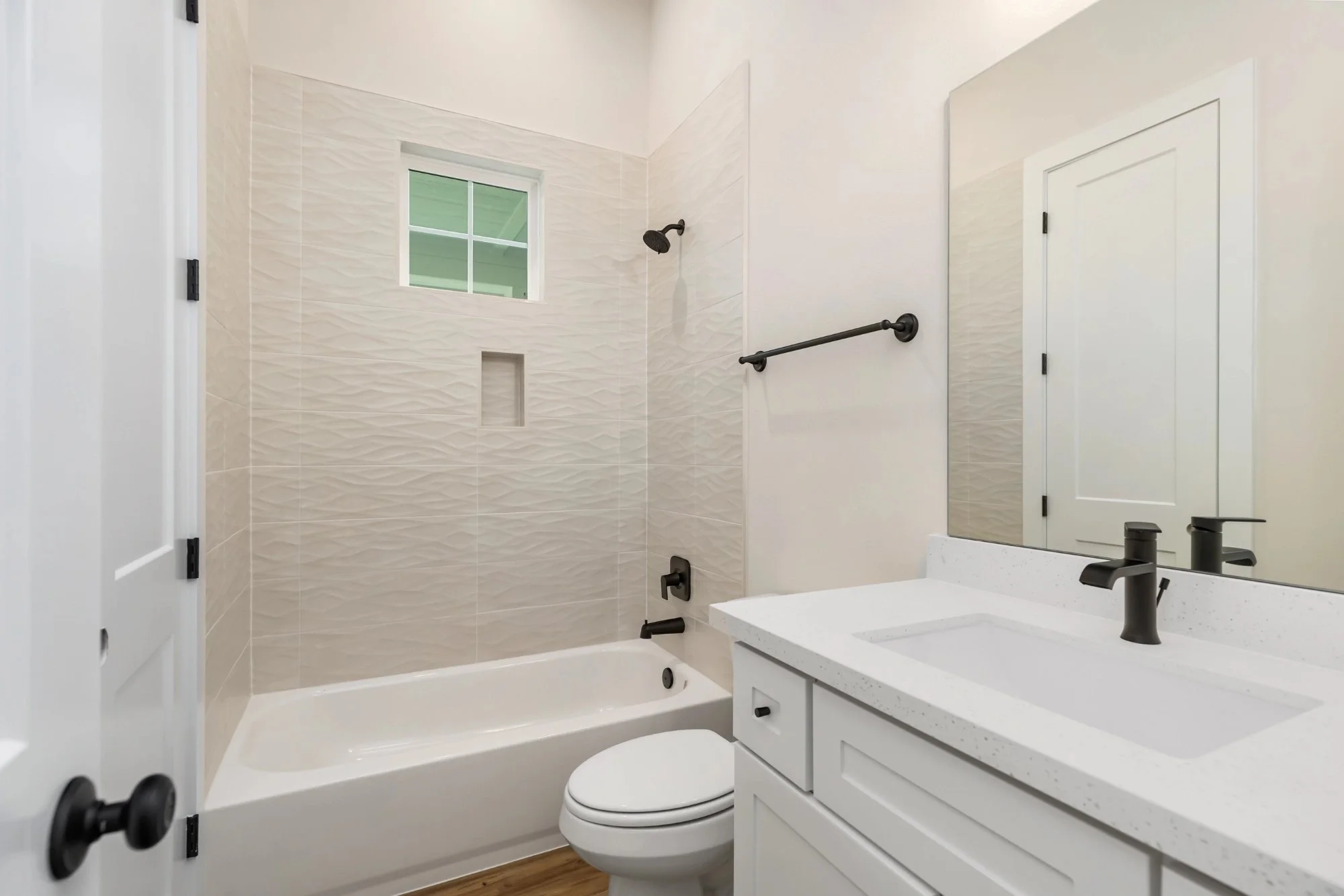 A clean, modern bathroom with a bathtub, toilet, white vanity with black fixtures, a mirror, a small window, and a black towel rack.