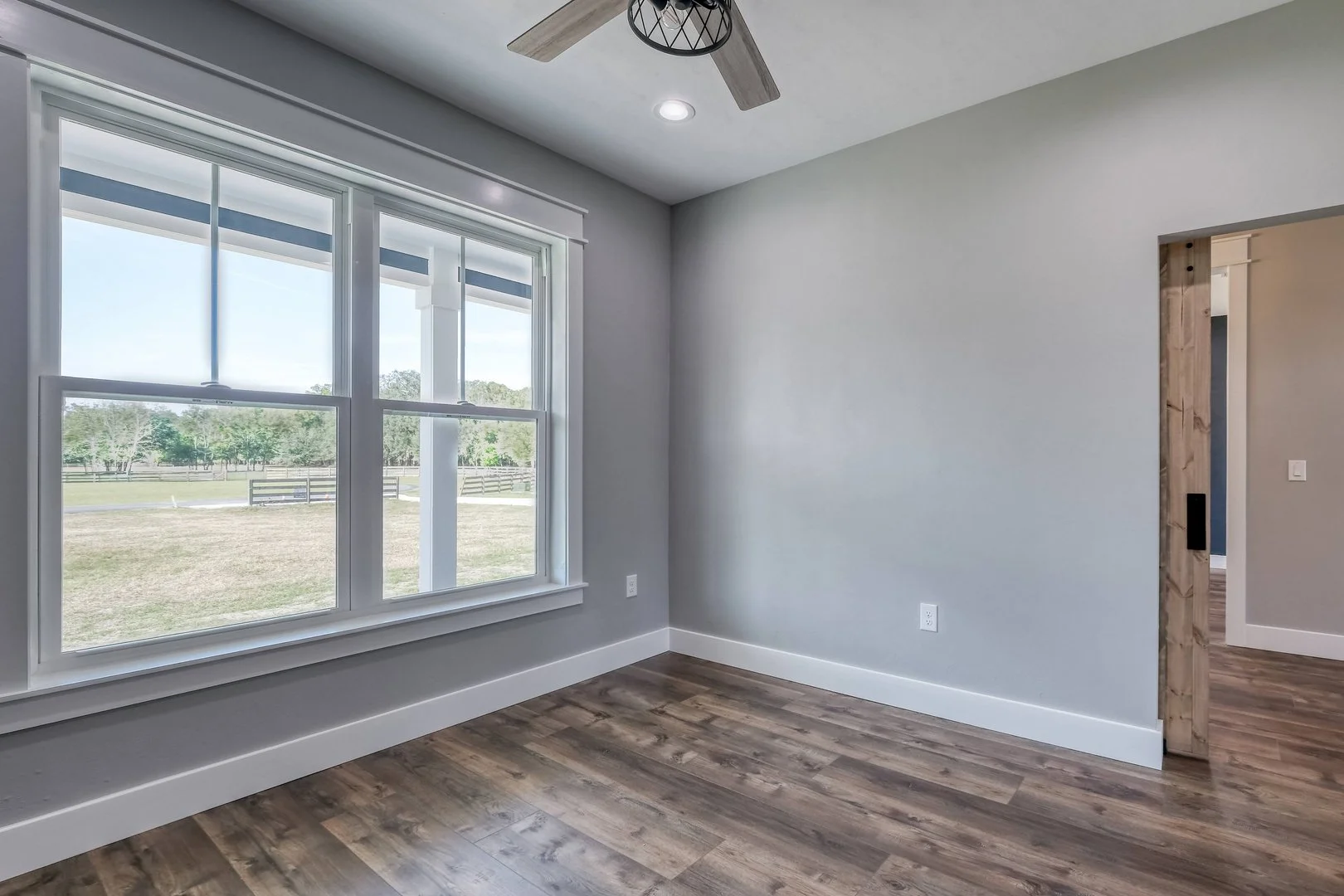 Empty room with large window showing a yard with trees, light gray walls, dark wood flooring, and a ceiling fan.