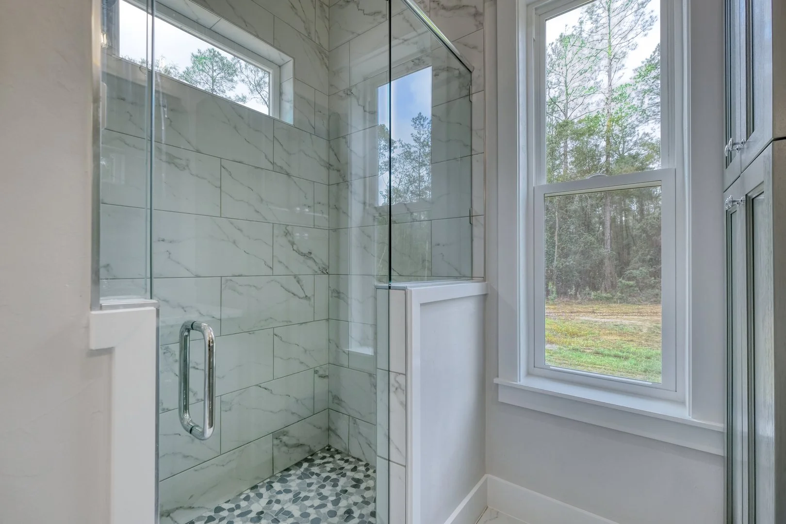 A modern bathroom shower with a glass door, white marble tiles, and a pebble stone floor, next to a large window showing an outdoor view of trees and sky.