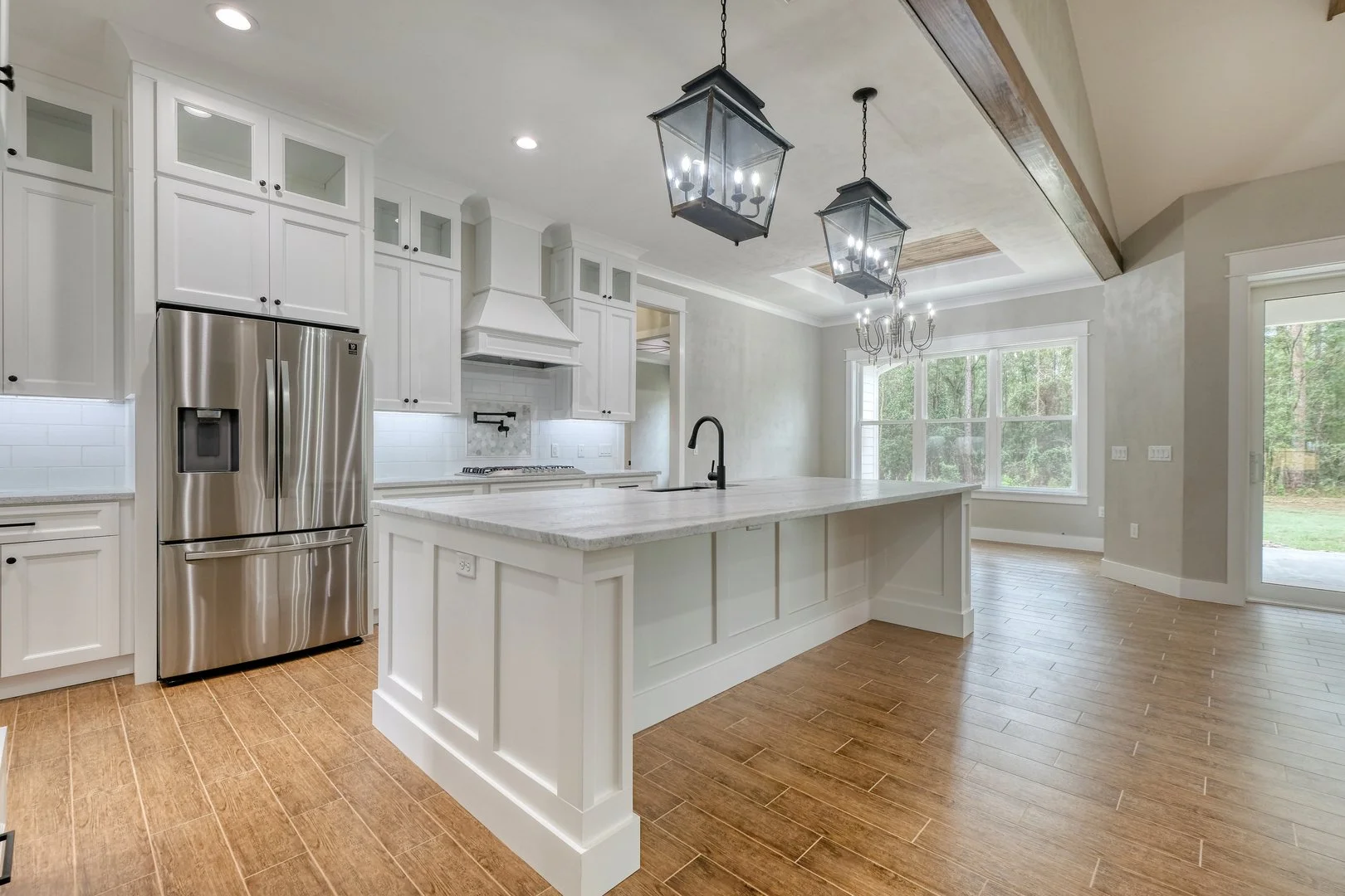 Modern kitchen with white cabinetry, stainless steel refrigerator, marble island, pendant lighting, and large windows with a view of trees.