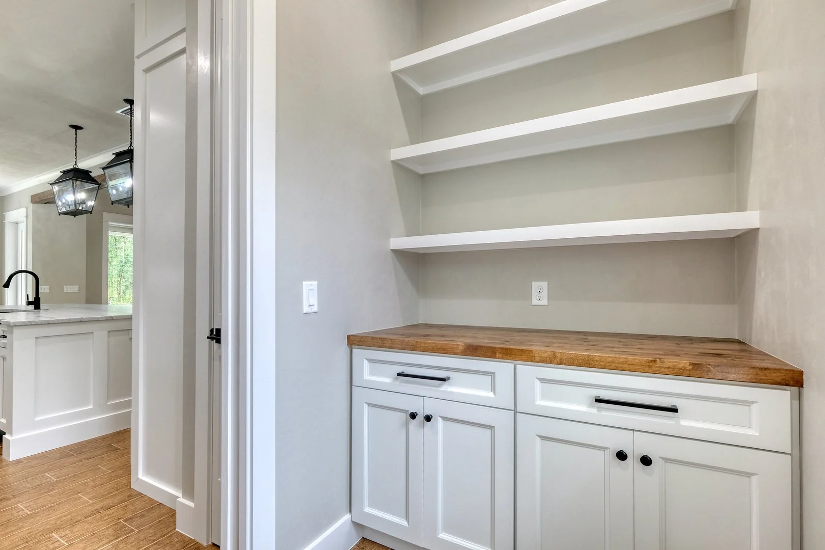 A corner of a kitchen with white cabinets, a wooden countertop, and three separated white floating shelves on a beige wall.