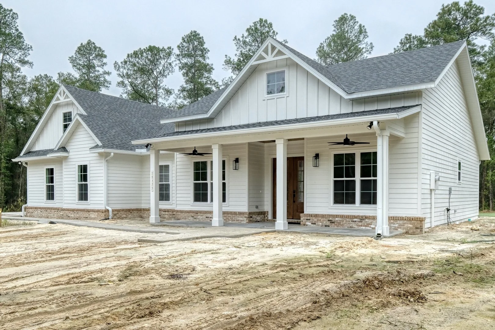 New white house with black roof, front porch with two white columns, ceiling fans, and multiple windows, surrounded by trees, on a dirt lot.