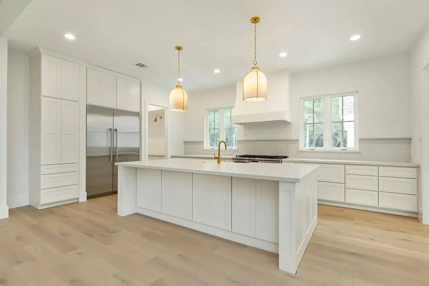 Modern, bright white kitchen with large island, light wood flooring, stainless steel refrigerator, white cabinets, and pendant lighting.
