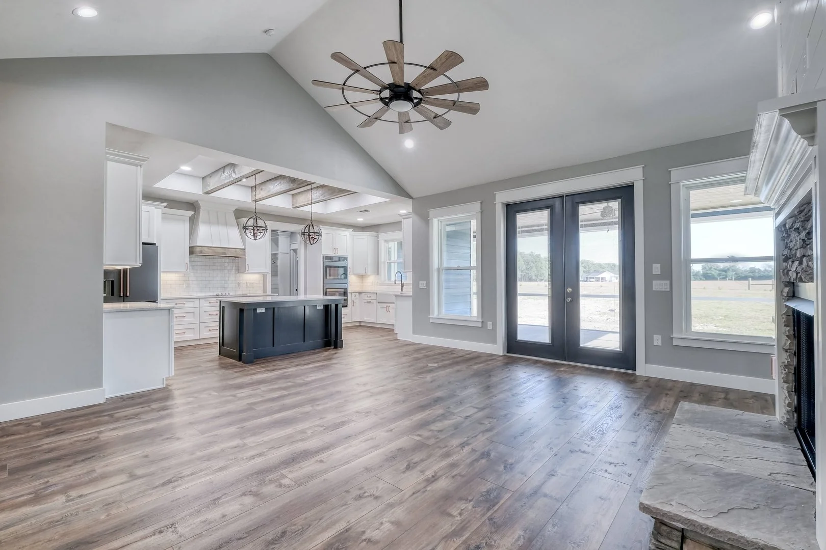 Empty living room with hardwood floors, a modern ceiling fan, and large windows with a view outside. The room connects to a white kitchen with an island, double ovens, and pendant lights, along with a set of glass double doors leading outside.