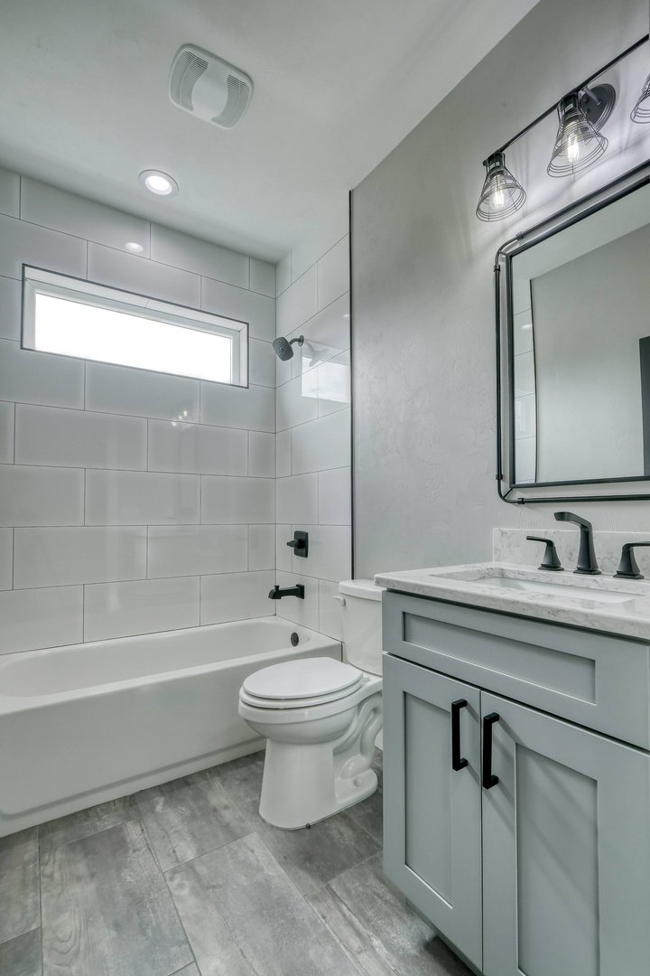 A modern bathroom featuring a white bathtub with a black faucet, a toilet, a light gray vanity with a marble countertop, a black faucet, a rectangular mirror, black light fixtures, a small horizontal window, and gray tiled flooring.