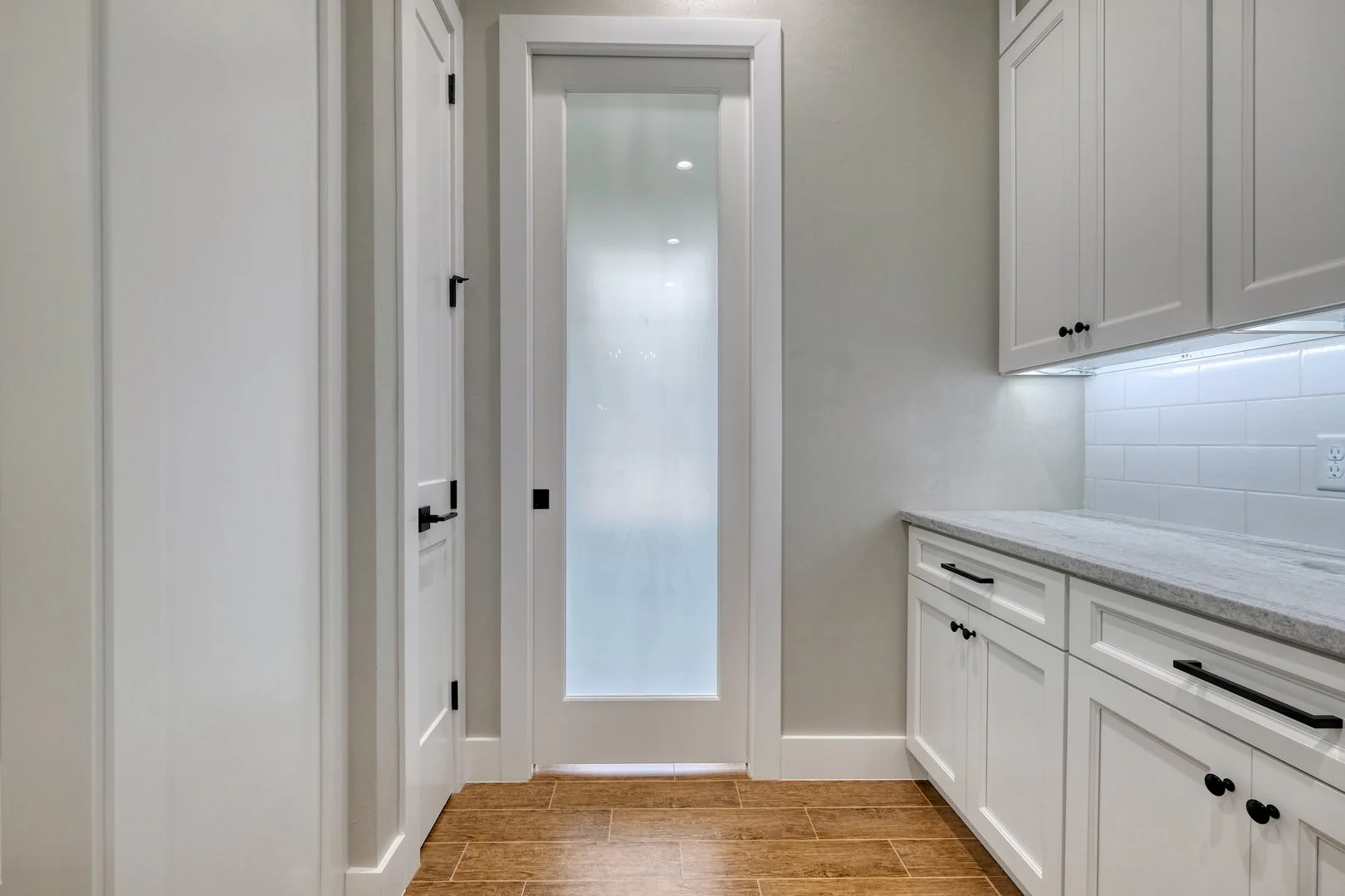 Interior view of a kitchen with white cabinets, a light gray countertop, a frosted glass door, and wooden flooring.
