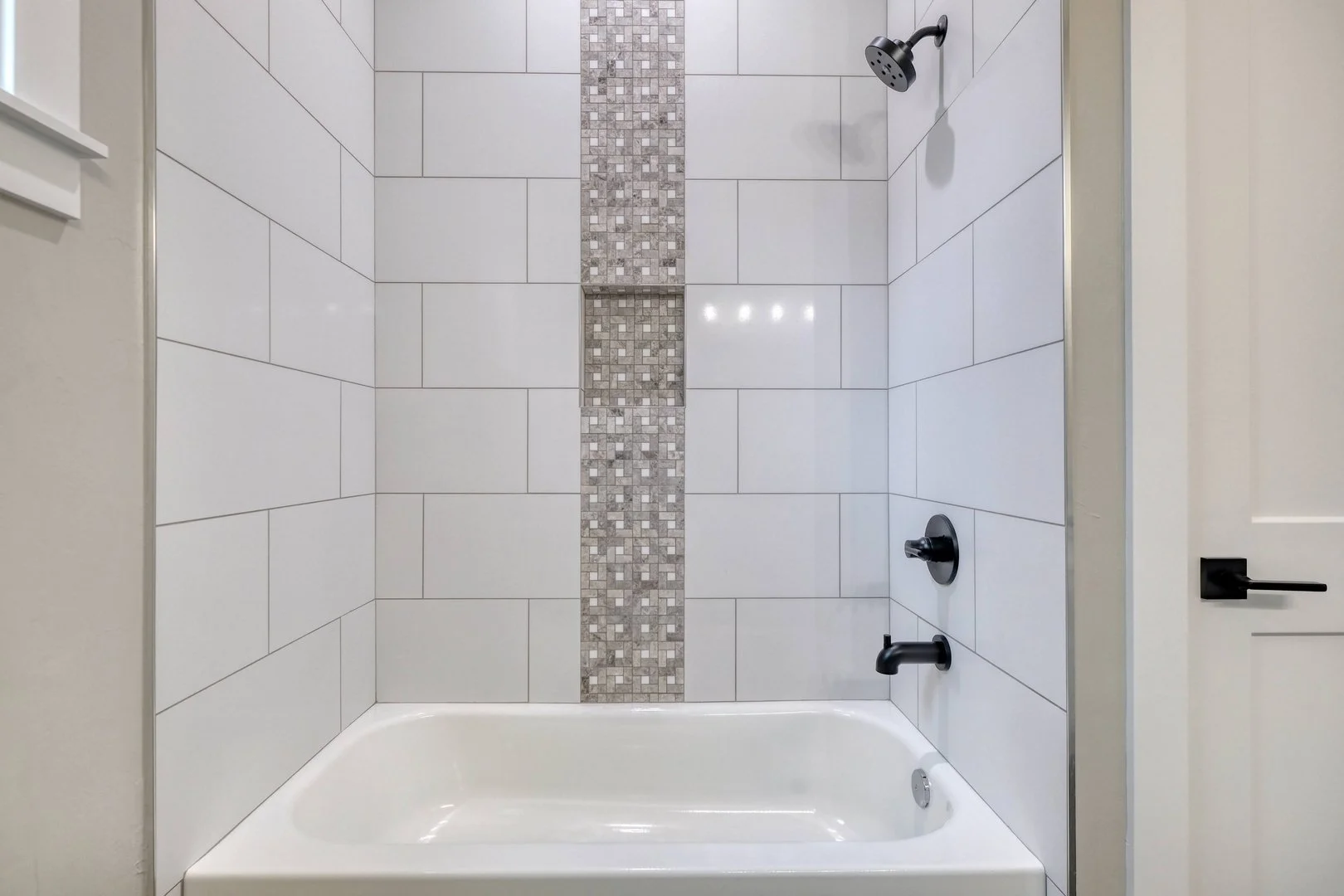 Modern walk-in shower with large white rectangular tiles and a vertical strip of small mosaic tiles in the center. The shower has black fixtures including a showerhead, faucet, and handle, with a white bathtub at the bottom. Beige wall to the side wi