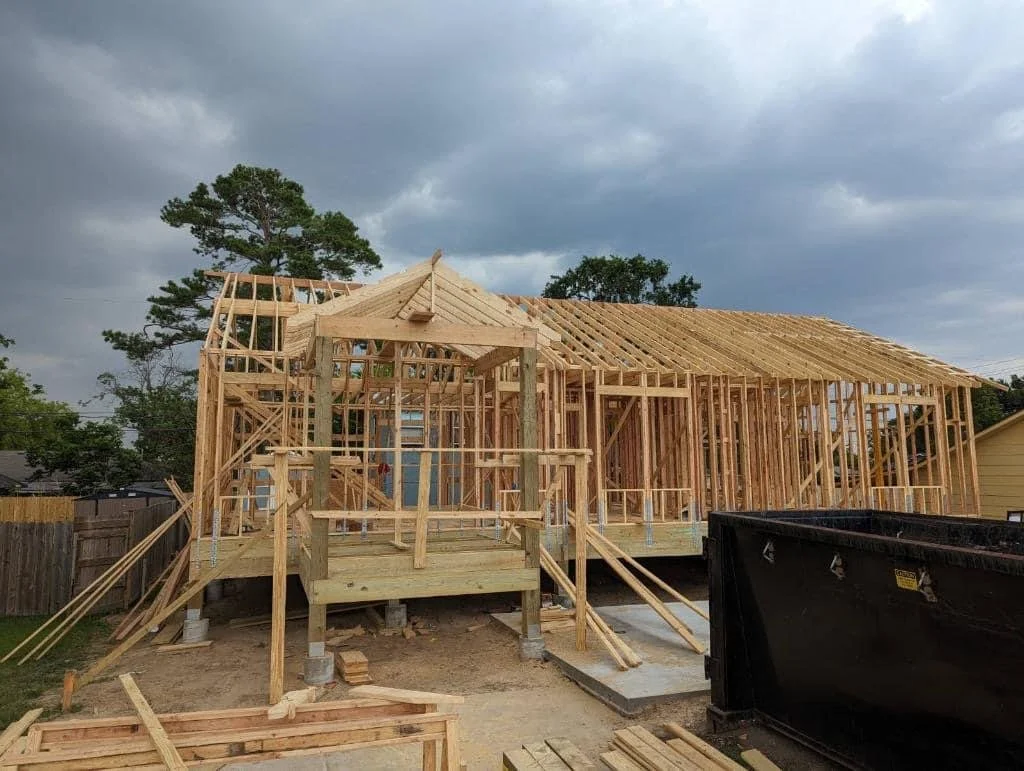 Construction site of a house frame with wooden beams and roof structure, cloudy sky in background.
