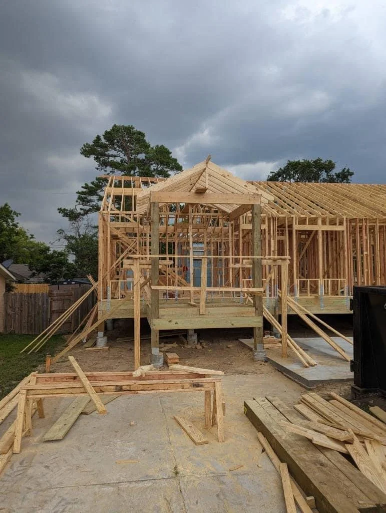 Construction site of a wooden house frame with a porch, set against a cloudy sky, surrounded by a wooden fence and some trees.