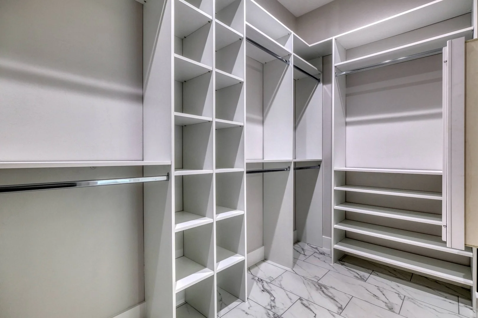 Empty walk-in closet with white shelves, drawers, and hanging rods, marble tile floor.