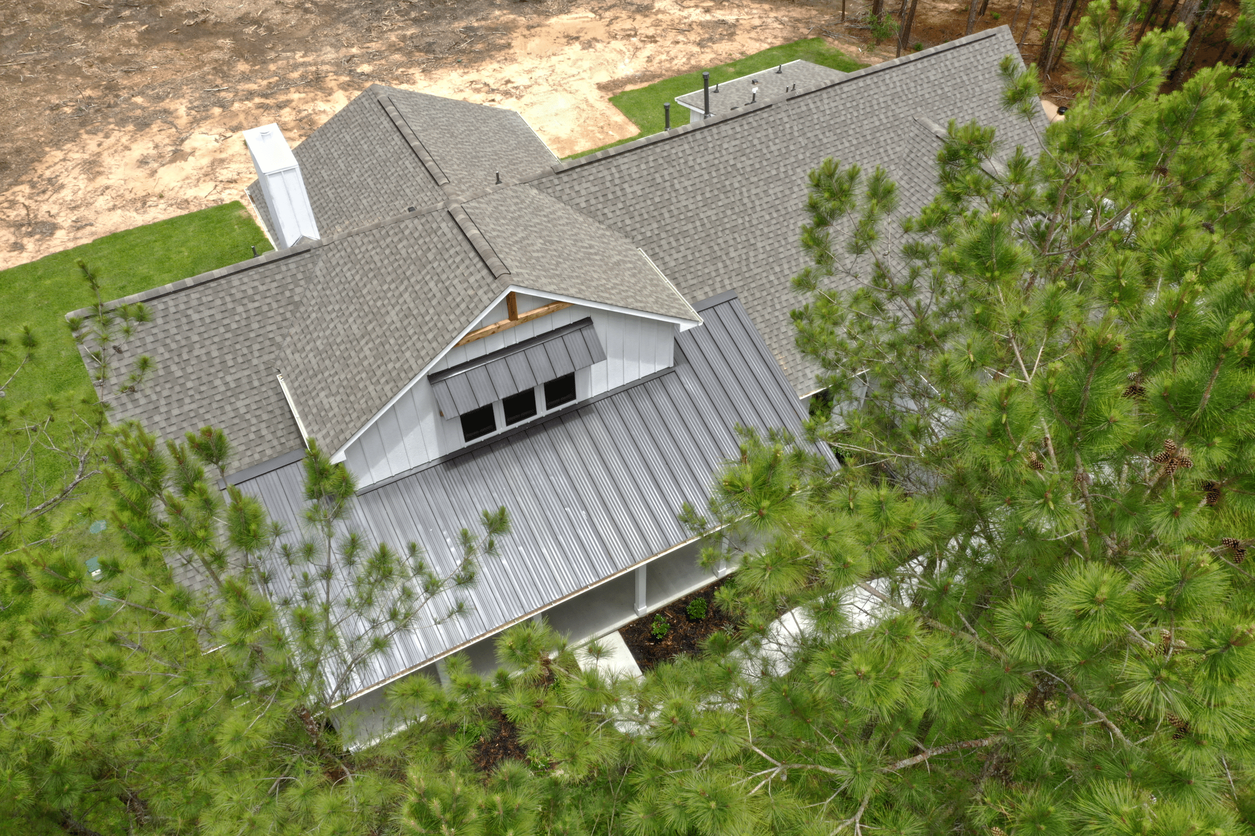 Aerial view of a house with gray roof, white siding, surrounded by green trees and a lawn, with a dirt yard nearby.