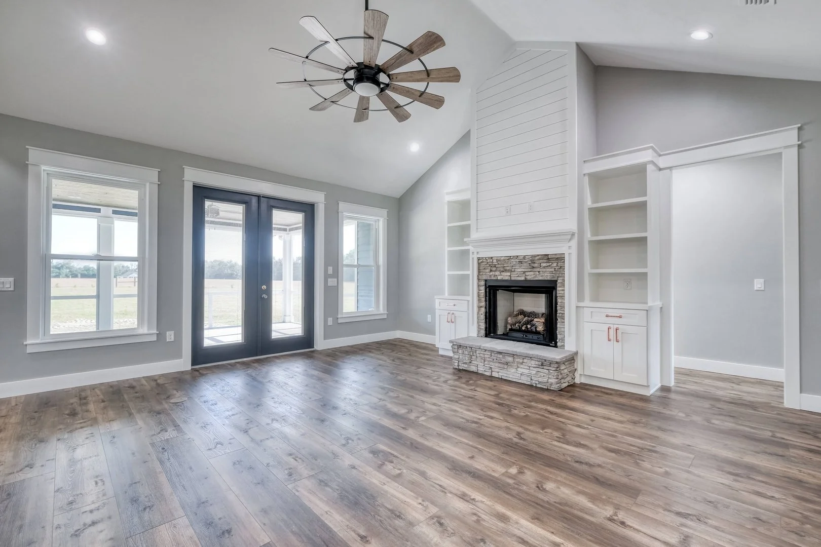 Empty living room with gray walls, hardwood floors, a stone fireplace with white wood trim, built-in shelves, three windows, and a black ceiling fan with wooden blades.