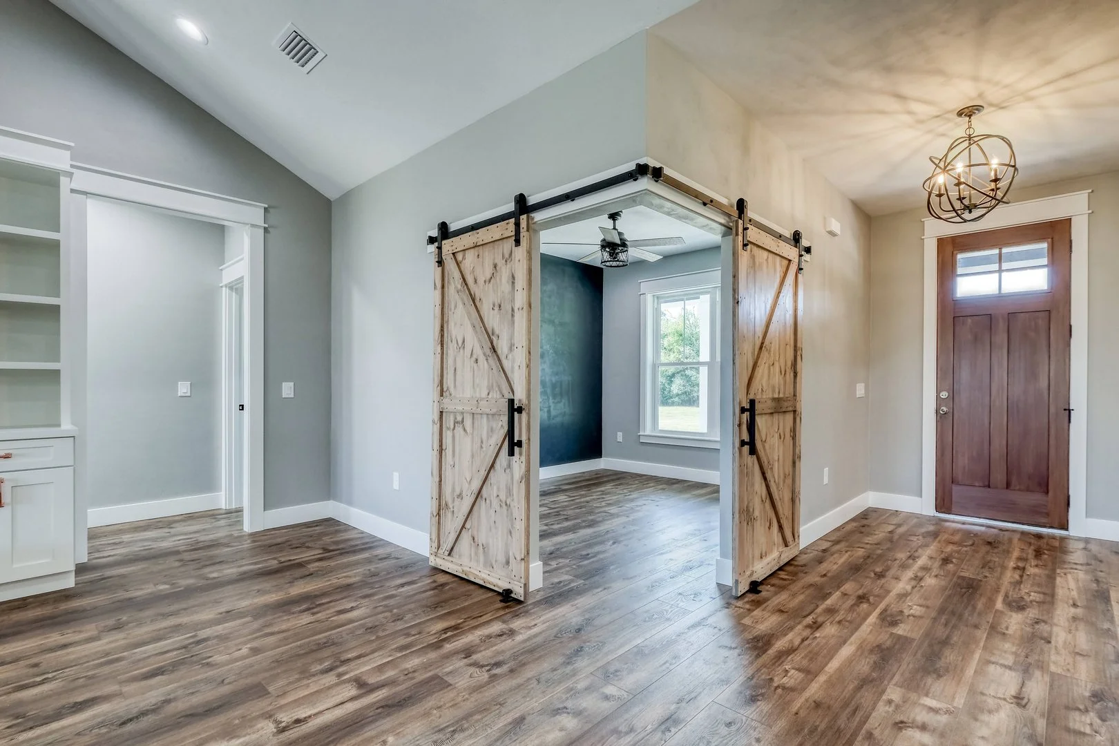 An interior room featuring wooden flooring, a barn door-style sliding door, a ceiling fan, a white built-in bookshelf, and a window showing greenery outside.