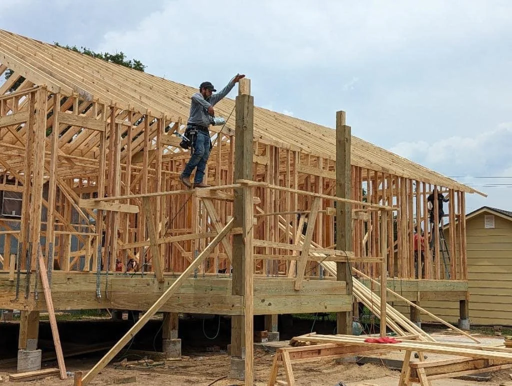 Workers constructing a wooden house framework with a pitched roof, using ladders and scaffolding at a construction site.