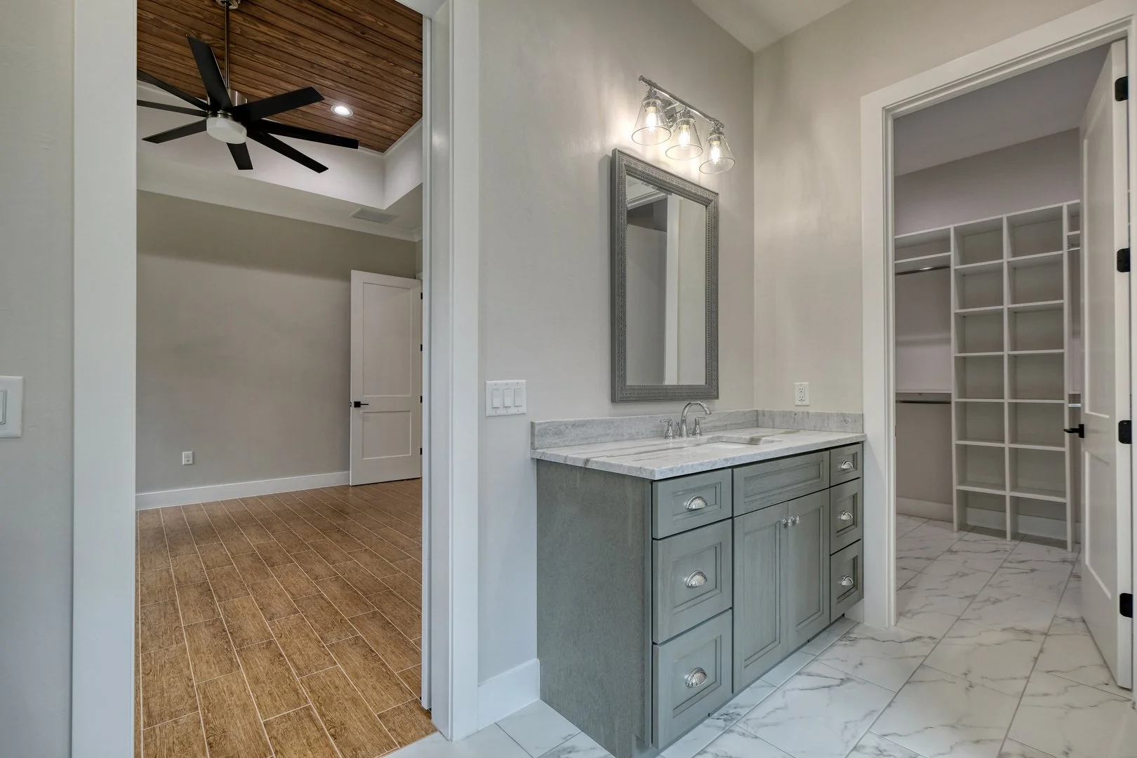 Bathroom with a gray vanity, marble countertop, mirror, and light fixture; walk-in closet with built-in shelves; wooden ceiling fan in adjacent room.