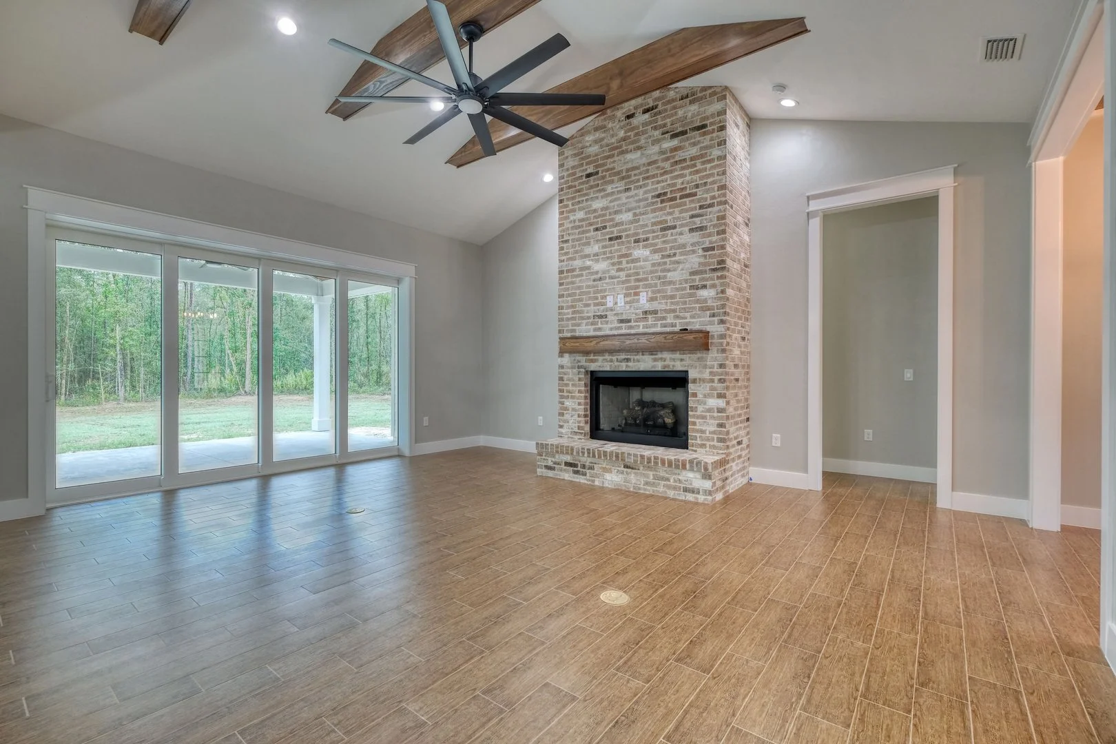 Living room with wood flooring, brick fireplace, large sliding glass doors leading outside, and a ceiling fan with wooden accents.