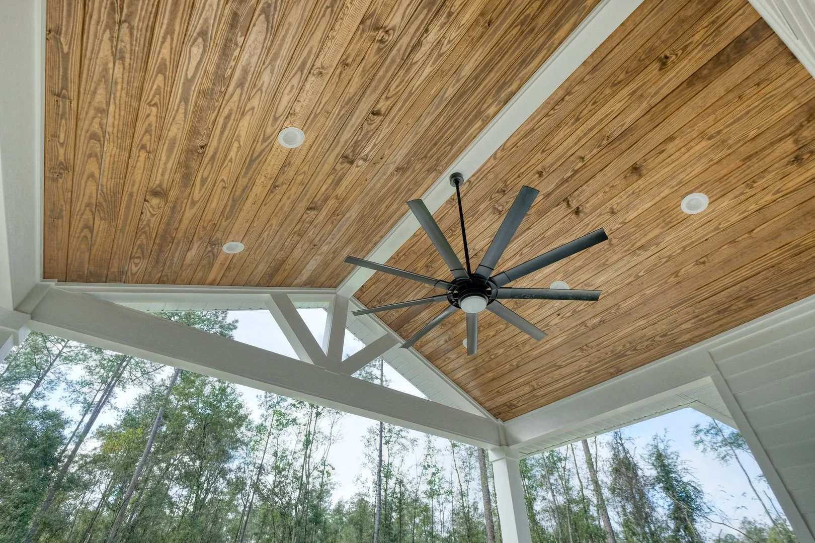 Ceiling with wooden planks, recessed lighting, and a modern black ceiling fan, with large windows that reveal a view of trees outside.