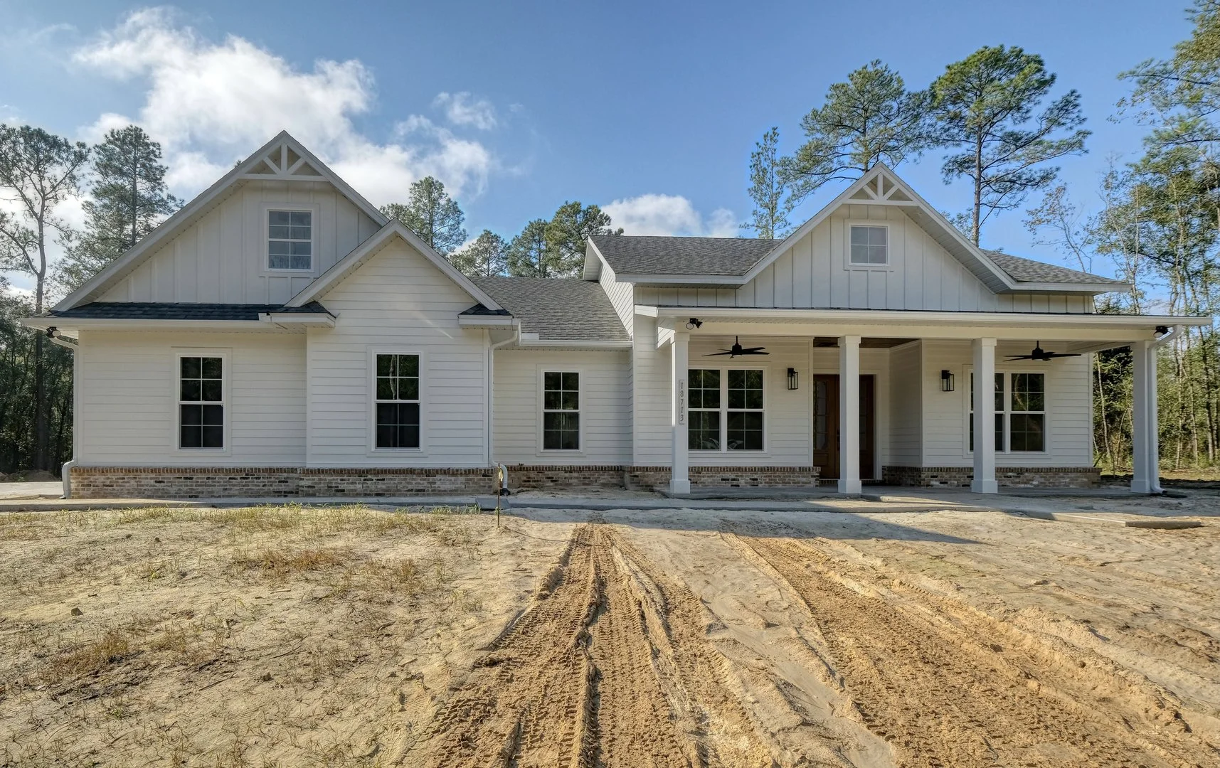 New white house with black roofing and a front porch, set against a blue sky with trees in the background, with dirt driveway in front.