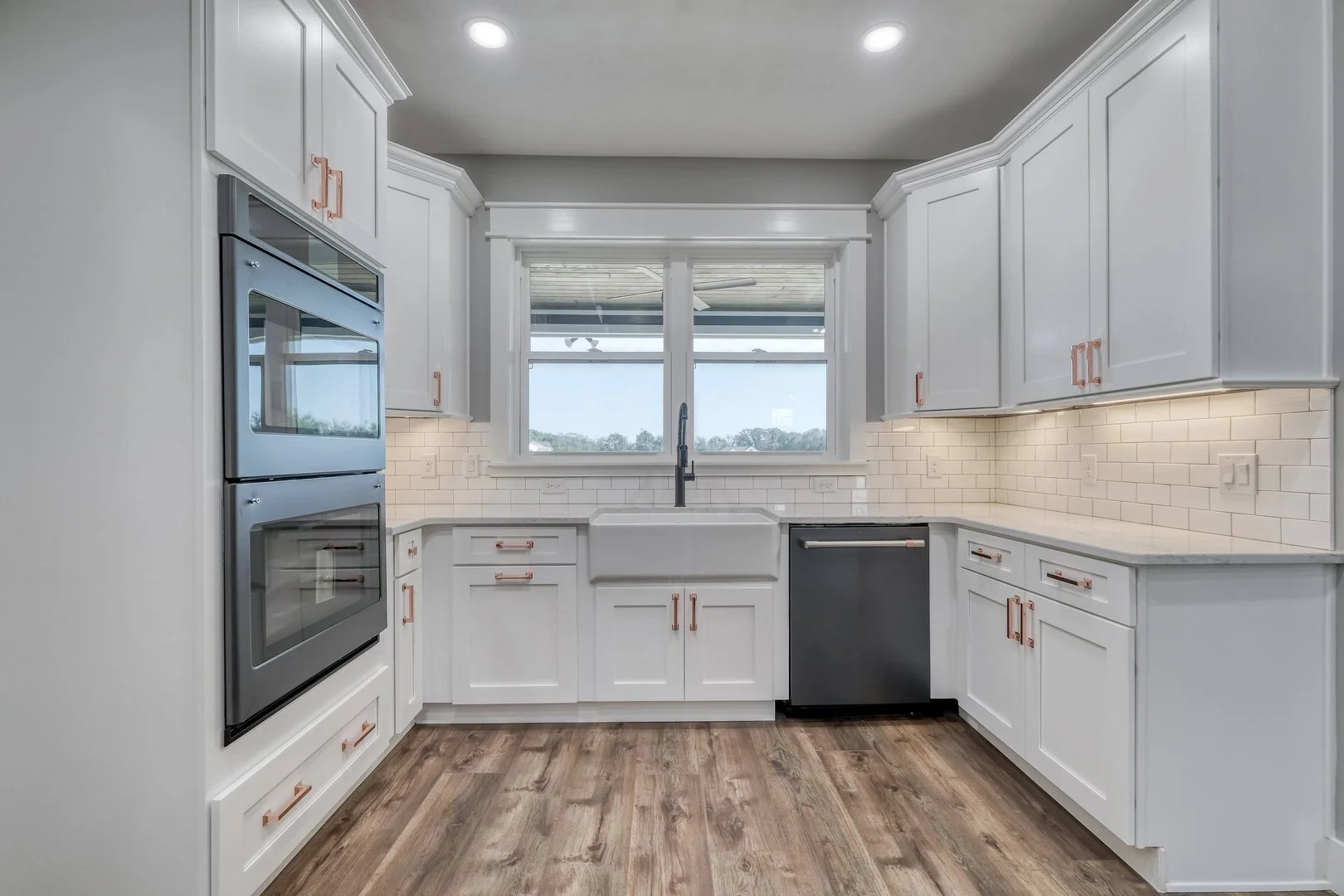 Modern kitchen with white cabinets, a large farmhouse sink, a window over the sink, gray double oven, and a black dishwasher, with wood flooring and white subway tile backsplash.