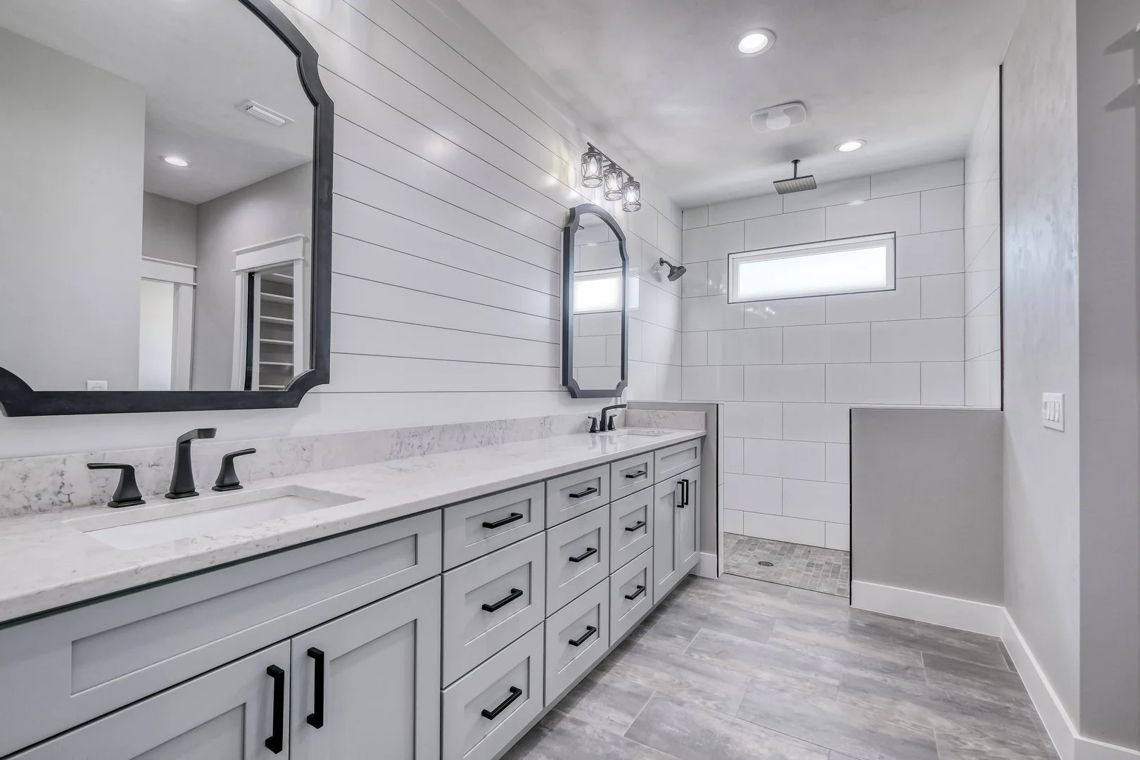 Modern bathroom with gray vanity, marble countertop, dual black faucets, large mirror, white shiplap wall, vanity lighting, gray flooring, walk-in shower with white tiles and a small window.
