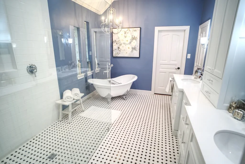 A bright bathroom with a blue wall, white vanity, and black-and-white tiled floor. Features a corner bathtub, towel rack, and pendant light fixture.