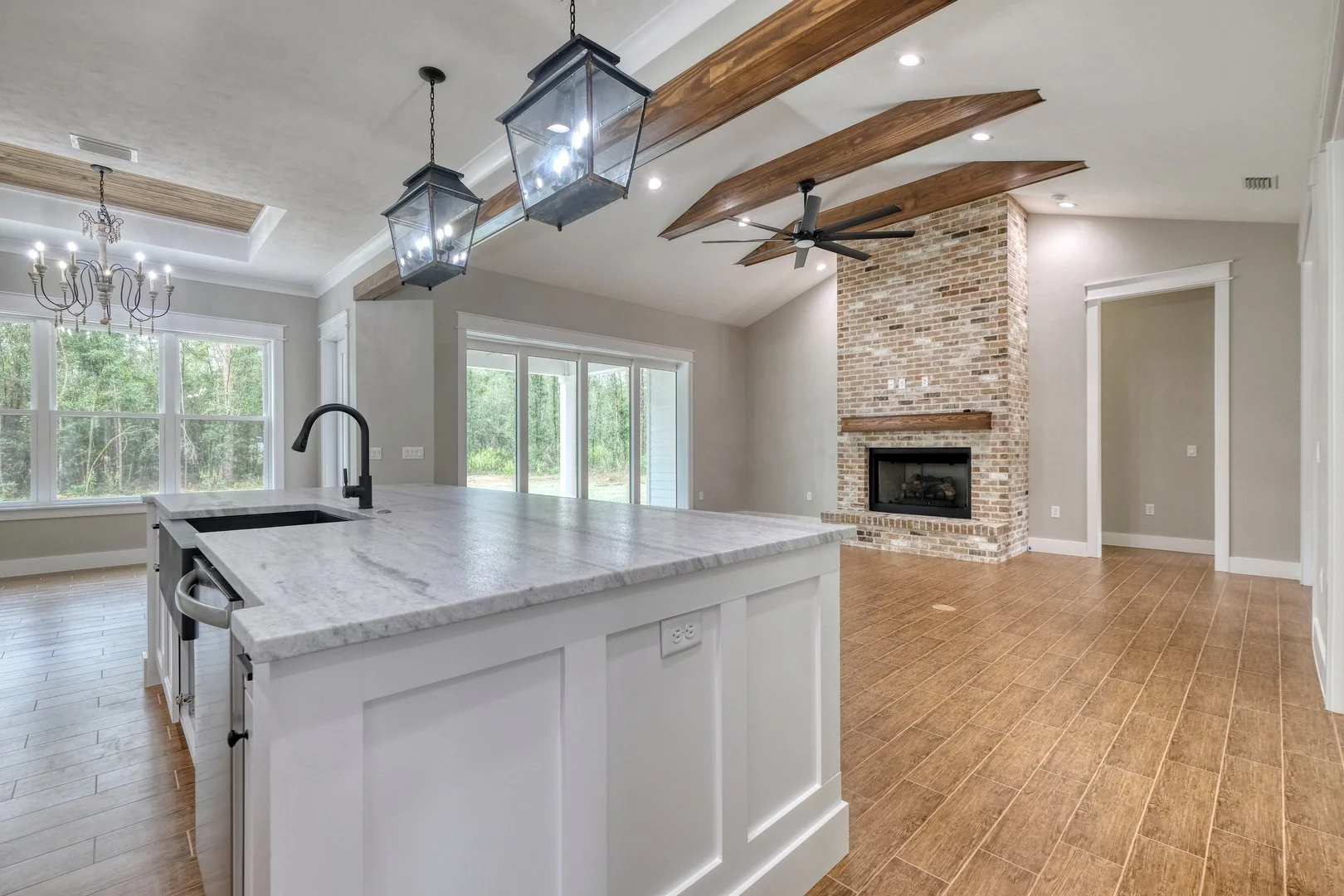 Open-concept living room and kitchen with hardwood floors, large windows, brick fireplace, wooden beams, ceiling fan, and modern lighting fixtures.