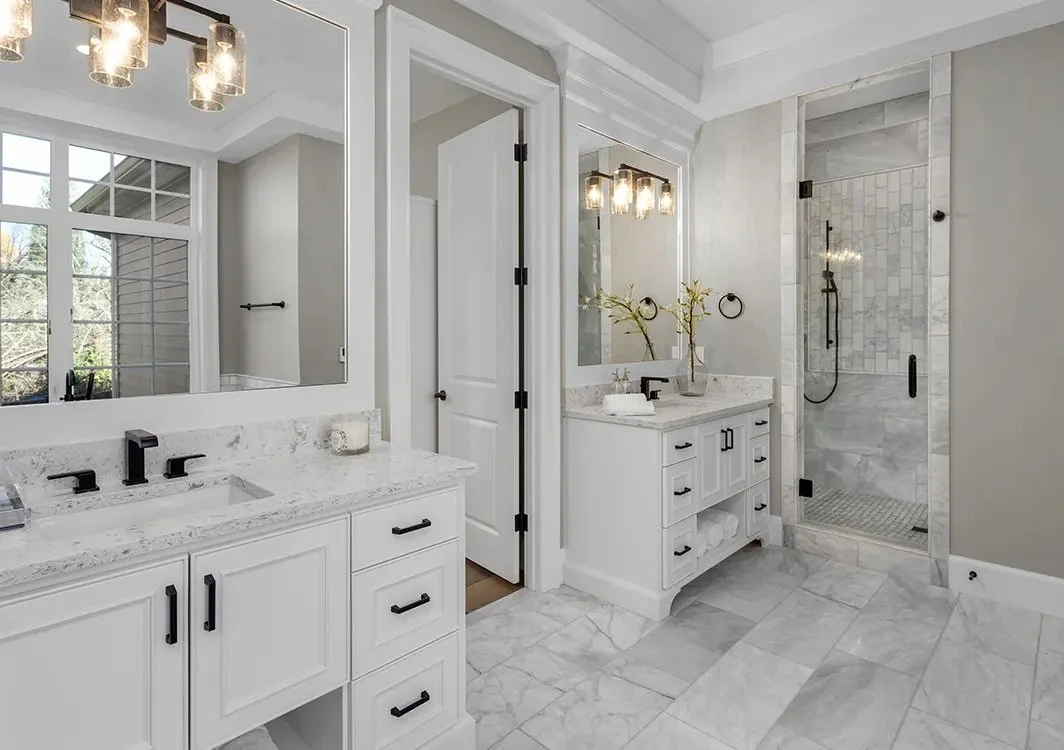 Modern bathroom with white cabinetry, marble countertops, two sinks, large mirror, and a walk-in glass shower with marble tiles.