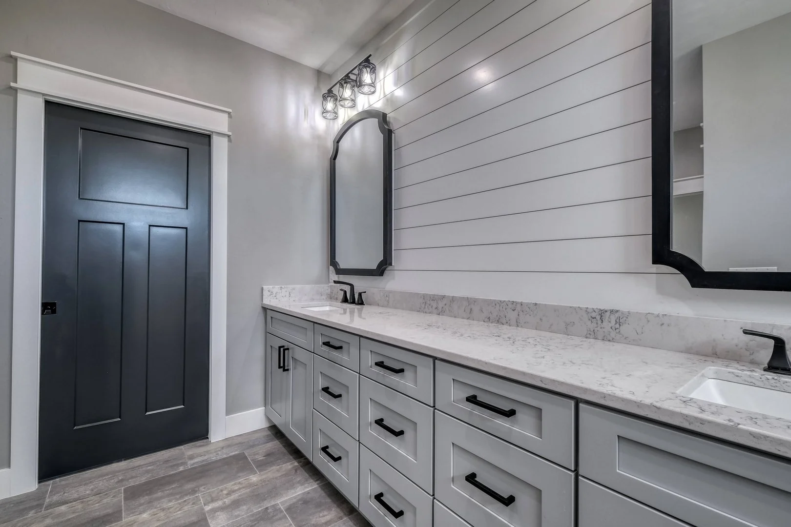 Modern bathroom vanity with gray cabinets, a marble countertop, two small sinks, black hardware, and two mirrors with black frames on a white shiplap wall, lit by a three-bulb light fixture.