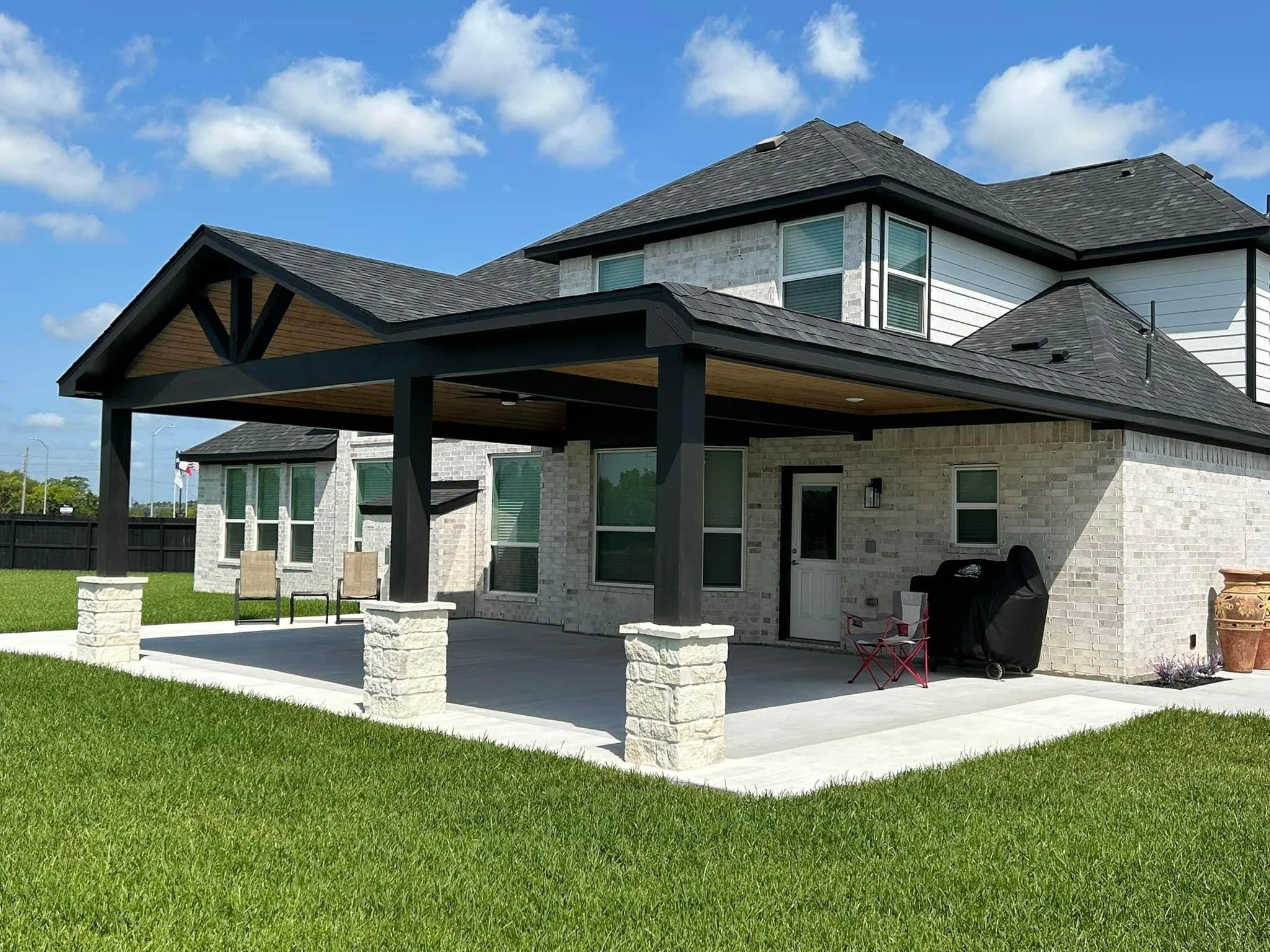Backyard of a modern two-story house with a covered patio, brick walls, multiple windows, and a well-maintained lawn under a partly cloudy sky.