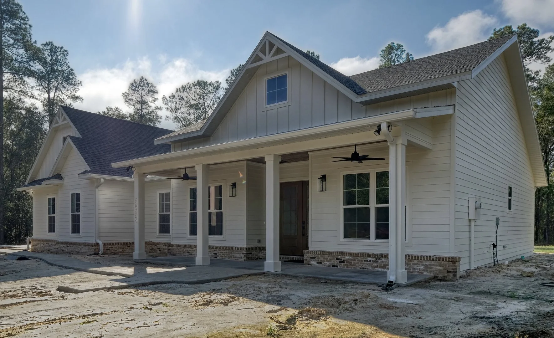 Newly constructed white house with a porch, multiple windows, a brick foundation, and a dark gray roof, surrounded by trees under a partly cloudy sky.