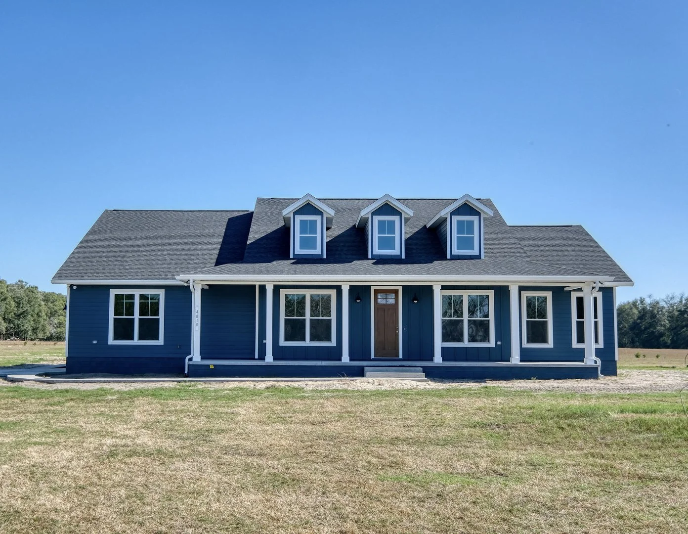 Front view of a new, blue, two-story house with a porch and three dormer windows under a clear blue sky.