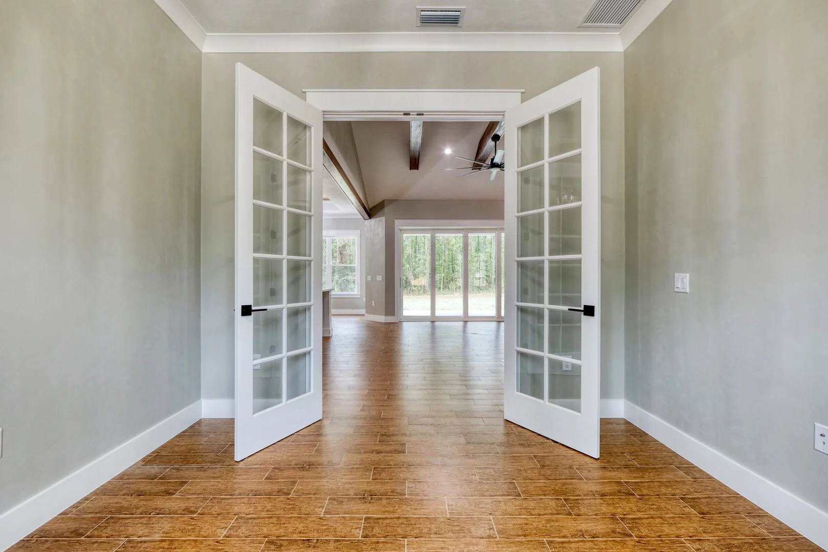 View through open glass-paneled double doors into a spacious living area with large windows and sliding glass doors leading outside, featuring wood flooring, beige walls, and a ceiling with exposed beams and a ceiling fan.