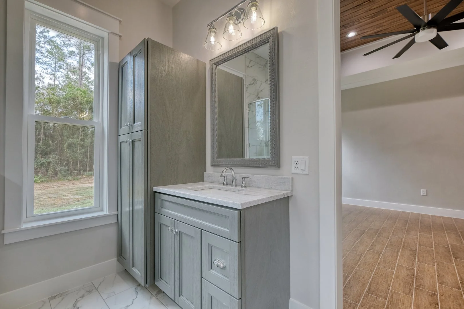 Bathroom with a vanity mirror, gray cabinetry, white marble countertop, and a window showing outdoor trees.
