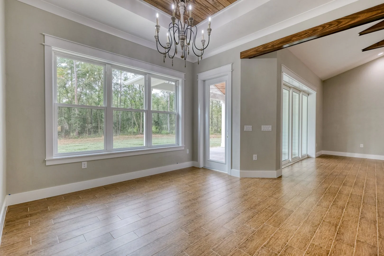 Empty living area with large window, hardwood flooring, light gray walls, white trim, and a chandelier hanging from a wooden ceiling section.